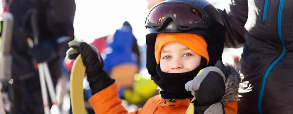 Enfant portant une cagoule enfant noire et orange sous un casque de ski avec des lunettes de protection, prêt pour les sports d'hiver et les activités en montagne.