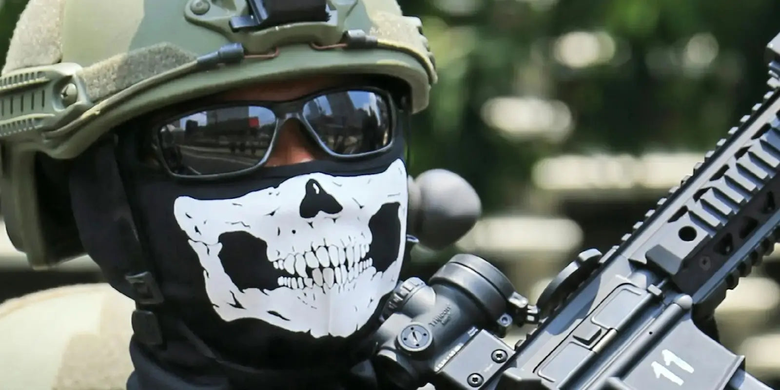 Militaire portant un casque tactique, des lunettes de soleil et une cagoule noire avec un motif de crâne blanc, dans un environnement militaire extérieur.