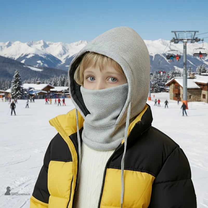 Bonnet cagoule enfant gris clair porté par un garçon sur une piste de ski avec chalets enneigés et télésiège en arrière-plan - Espace Cagoules