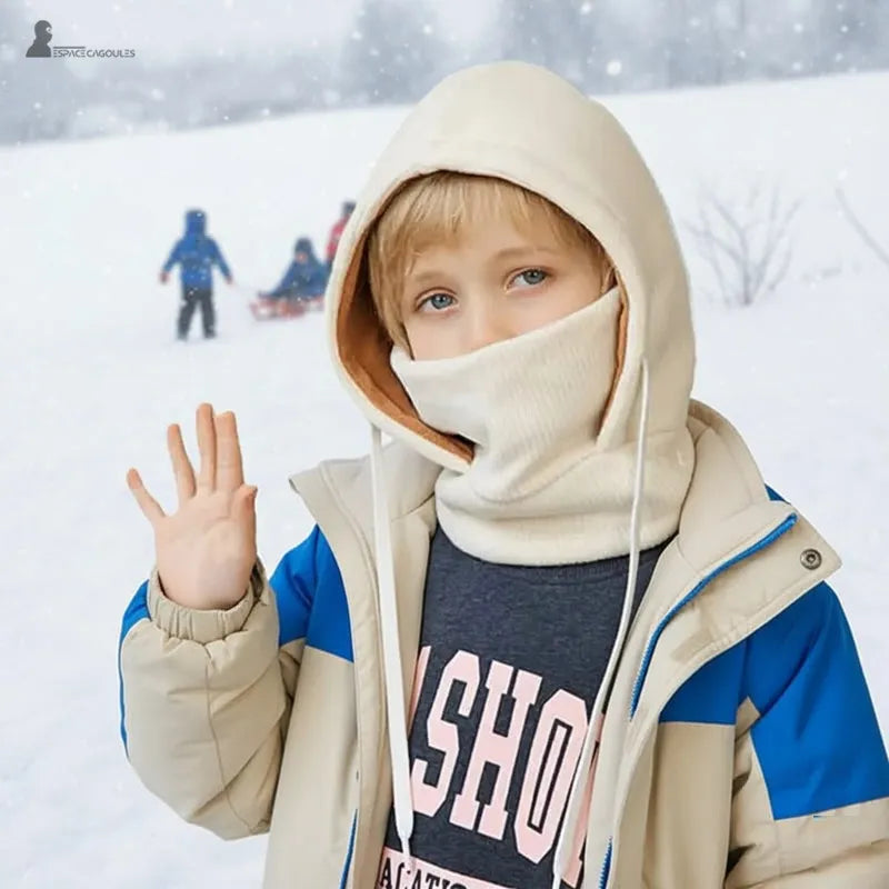 Bonnet cagoule enfant beige et bleu porté par un garçon saluant sous la neige pendant une journée d’hiver - Espace Cagoules