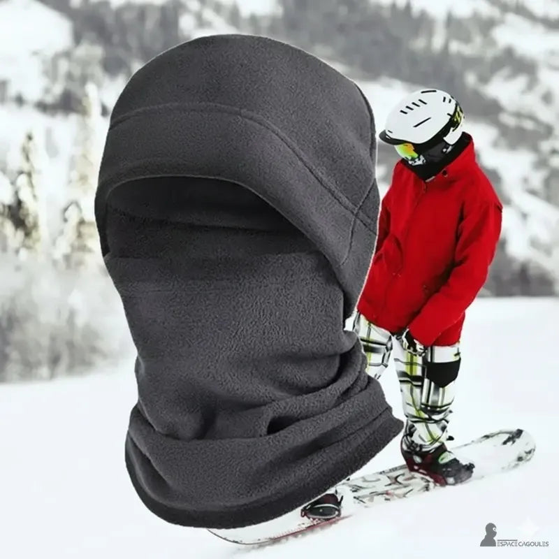 Bonnet cagoule gray porté sur fond enneigé avec un snowboarder en arrière-plan, protection idéale pour sports d’hiver Espace Cagoules