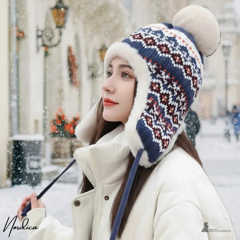 Blaue Hapka-Mütze, die von einer Frau in einem weißen Mantel auf einer verschneiten Straße getragen wird, Mütze mit Bommel und beigem Fell