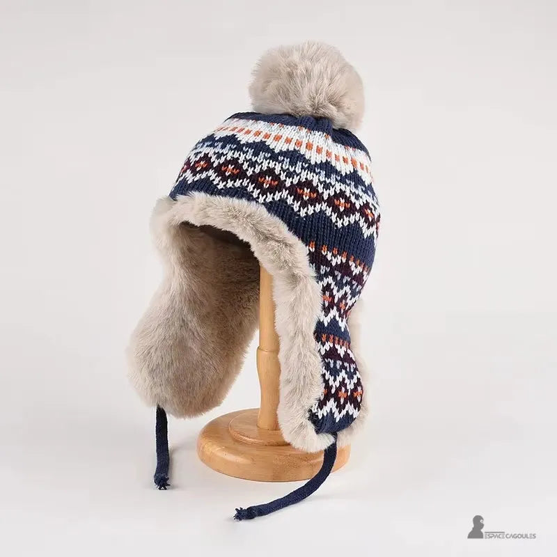 Blaue Hapka-Mütze mit geometrischen Mustern mit Bommel und beigem Kunstpelz-Interieur, präsentiert auf einem Holzständer