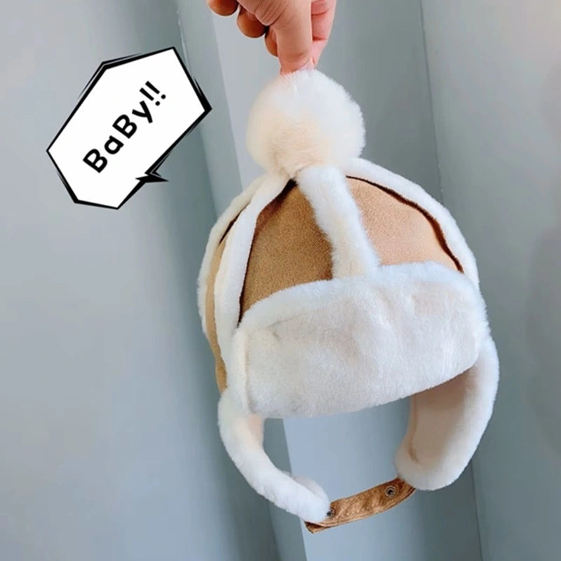 Children's Chapka beanie in beige and white fabric with a faux fur lining, earflaps and a wool pompom, held by a hand in a bright indoor environment.