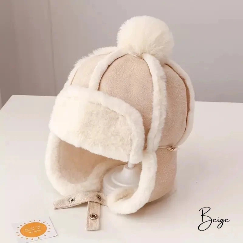 Close-up of a Chapka Enfant hat in beige fabric with visible seams, decorated with a fluffy pompom and a white faux fur interior, placed on a light wood table.