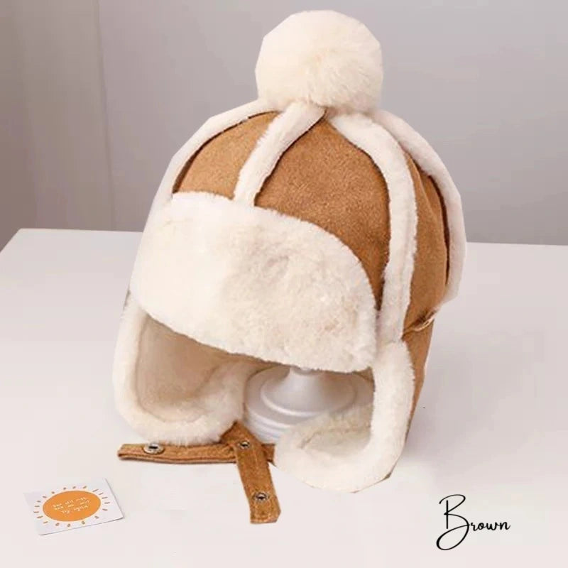 Brown Children's Chapka Hat with white faux fur interior, wool pompom and fold-over ear flaps, placed on a white support in a minimalist frame.