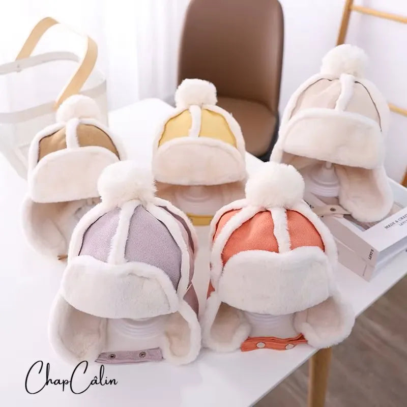 Children's chapka hat in soft faux fur with pompom, available in several pastel and bright colors, placed on a modern table