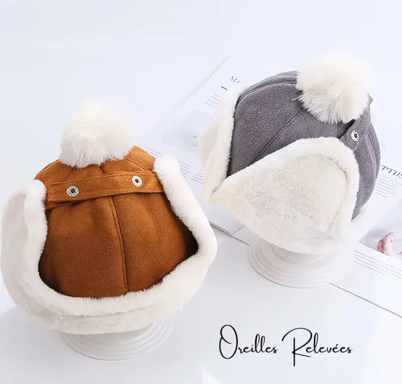 Children's Chapka beanies in 2 different shades, kakhi and grey and with a white faux fur interior and a wool pompom, carefully arranged for an elegant presentation.