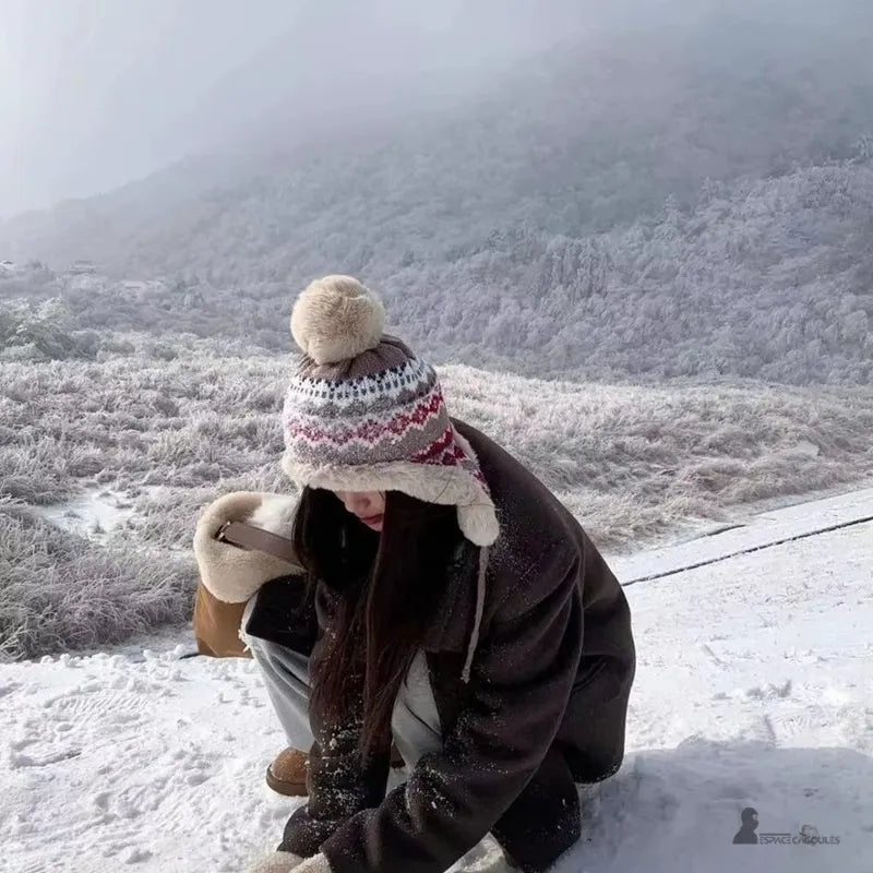 Beige und rote Hapka-Mütze, die von einer Frau getragen wird, die in einer verschneiten Berglandschaft kauert, Mütze mit Bommel und Ohrenklappen