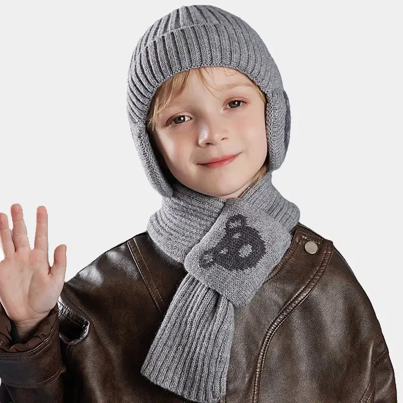 Enfant souriant portant un ensemble bonnet Ă©charpe enfant en tricot gris, avec un motif d'ours sur l'Ă©charpe, vĂȘtu d'une veste en cuir marron, sur un fond neutre.