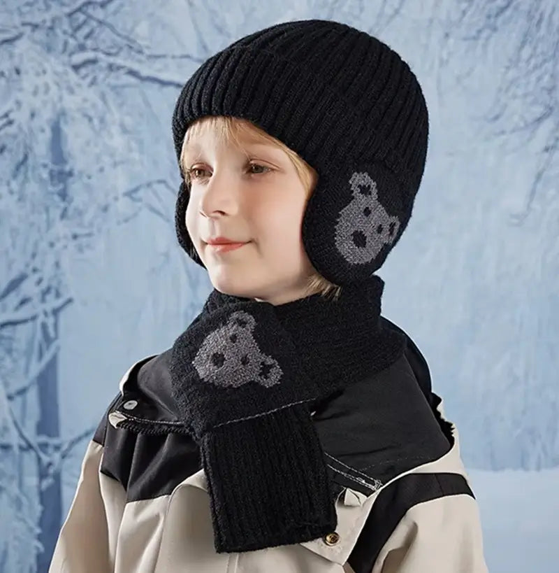 Garçon portant un ensemble bonnet Ă©charpe enfant en tricot noir avec des motifs d'ours gris, vĂȘtu d'une veste d'hiver noire et beige, posant devant un fond givrĂ© Ă©voquant un paysage hivernal.