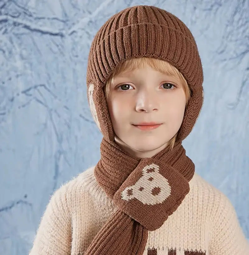 Garçon portant un ensemble bonnet Ă©charpe enfant en tricot marron avec des motifs d'ours beige, vĂȘtu d'un pull clair texturĂ©, posĂ© devant un fond bleu glacĂ© Ă©voquant l'hiver.
