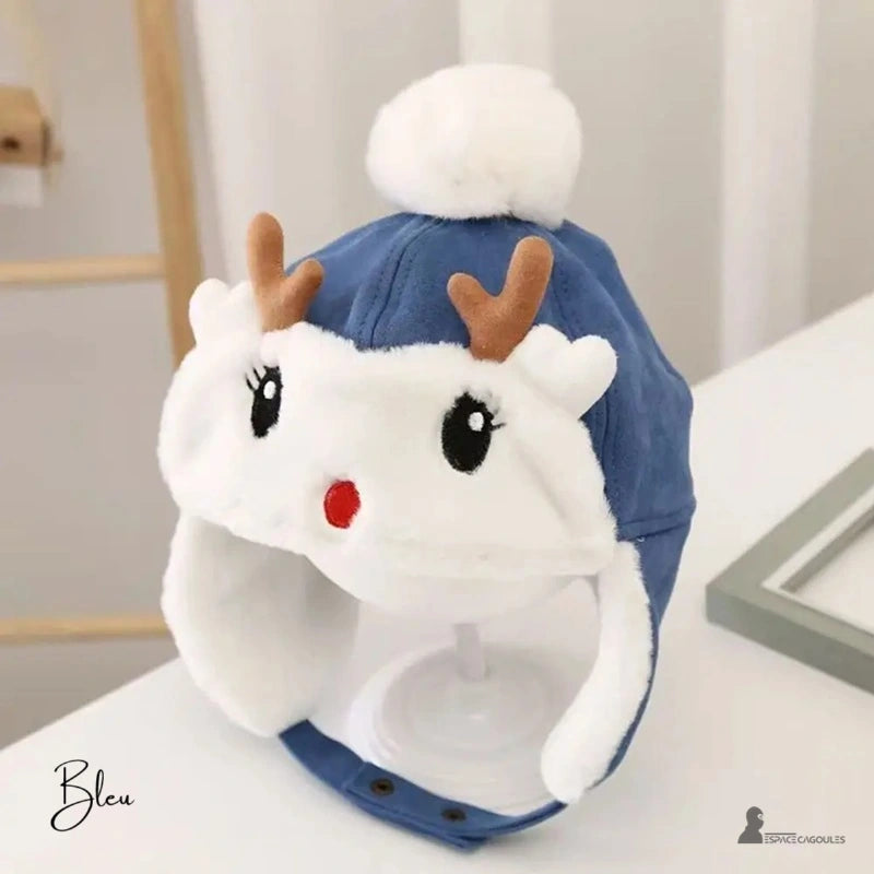Blue fleece reindeer children's hat with brown ears and white pompom, placed on a support indoors