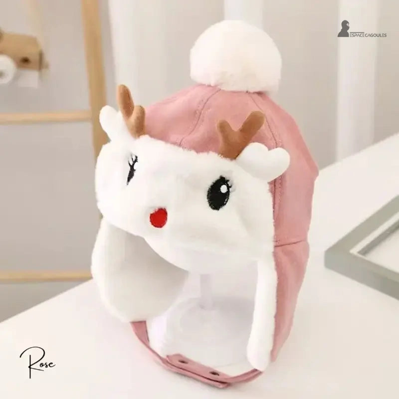 Pink fleece reindeer children's hat with brown ears and white pompom, placed on a support indoors