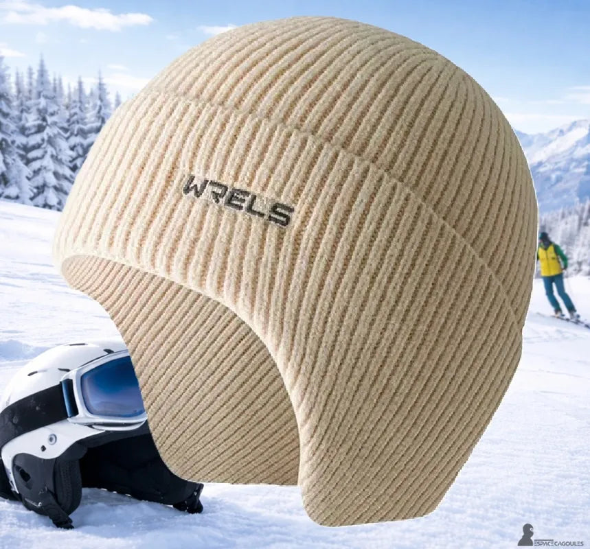 Bonnet sous casque ski en maille tricotée beige avec cache-oreilles intégré, vue de profil, posé devant un casque de ski sur neige en environnement montagne hivernal - Espace Cagoules