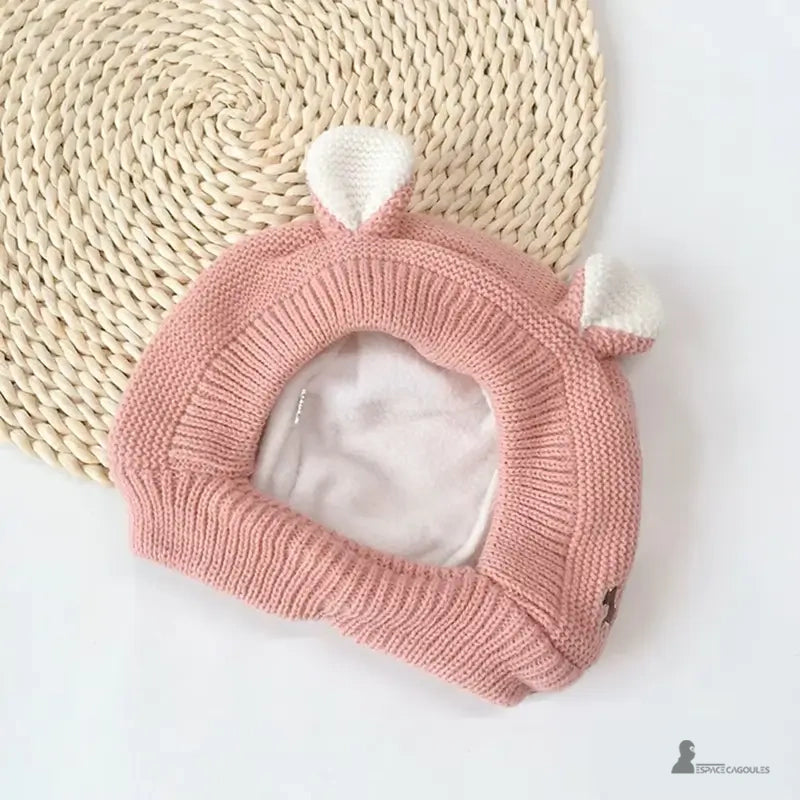 Cagoule Bébé 6 à 36 mois tricot rose avec oreilles blanches, posée à plat sur tapis rond en fibres tressées, doublure polaire douce visible