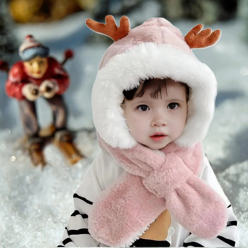Bébé avec une cagoule bébé fille blanche et rose en fausse fourrure, ornée de petites cornes de cerf, avec une écharpe intégrée croisée sous le menton. L'enfant est en extérieur sur un fond enneigé, avec une décoration hivernale en arrière-plan.