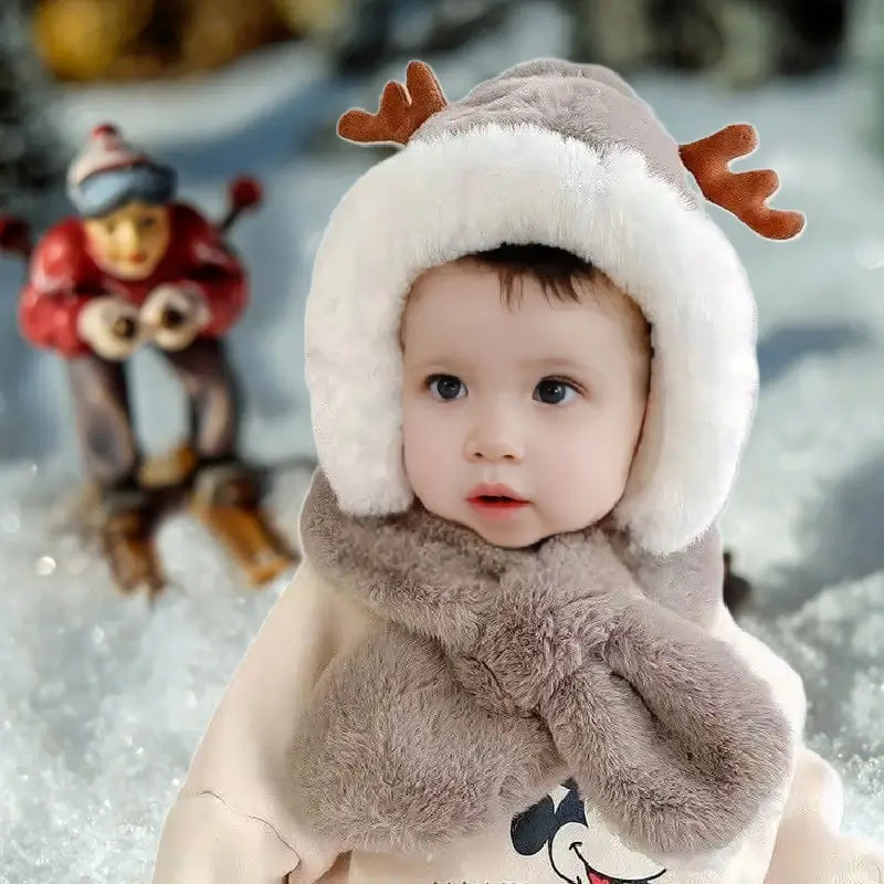 Braune Fleece-Sturmhaube für Jungen mit Rentierohren und flauschigem weißem Futter, das Kopf, Hals und Ohren bedeckt. Baby in einem beigen Wintermantel in einer verschneiten Umgebung mit einem Miniaturskifahrer im Hintergrund.