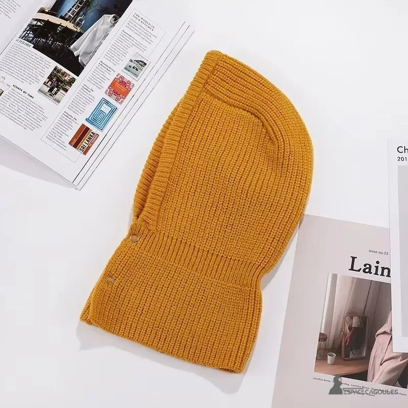 Cagoule Chaude pour Femme jaune moutarde tricotée, présentée à plat sur une table blanche avec des magazines autour.