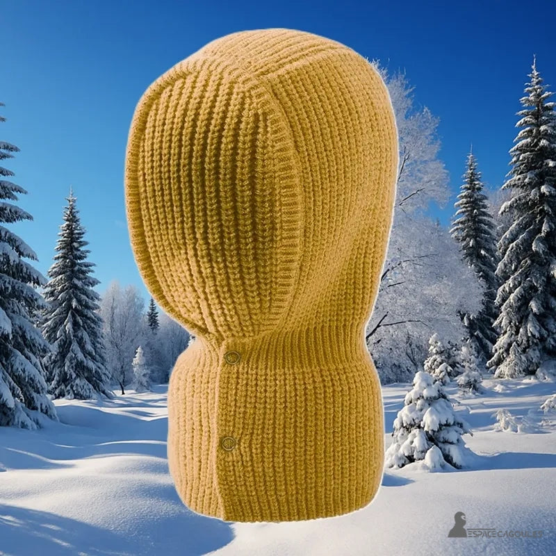 Cagoule Chaude pour Femme tricotée jaune moutarde avec boutons visibles, posée dans un décor hivernal enneigé avec sapins givrés.