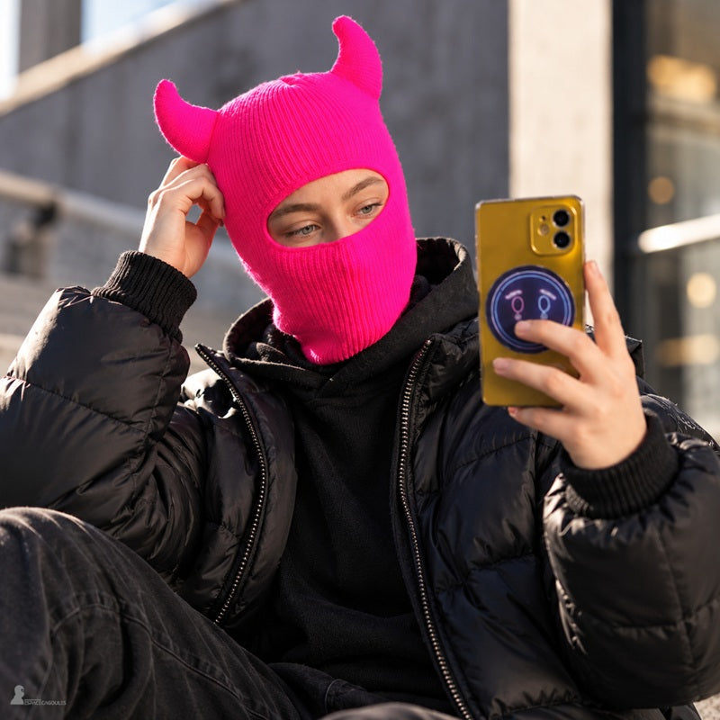 Femme en doudoune noire portant la cagoule diable rose fluo à cornes, selfie en ville - Espace Cagoules