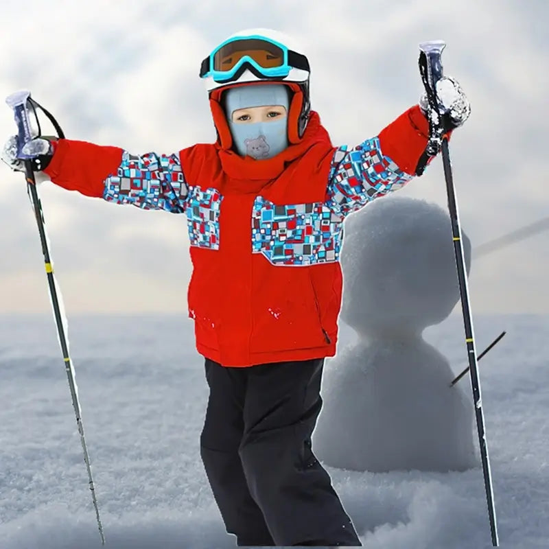 3-Ärigt barn som bÀr en rosa balaklava med ett dinosauriemönster, klÀdd i en röd jacka, njuter av snön med skidstavar i vintermiljö.