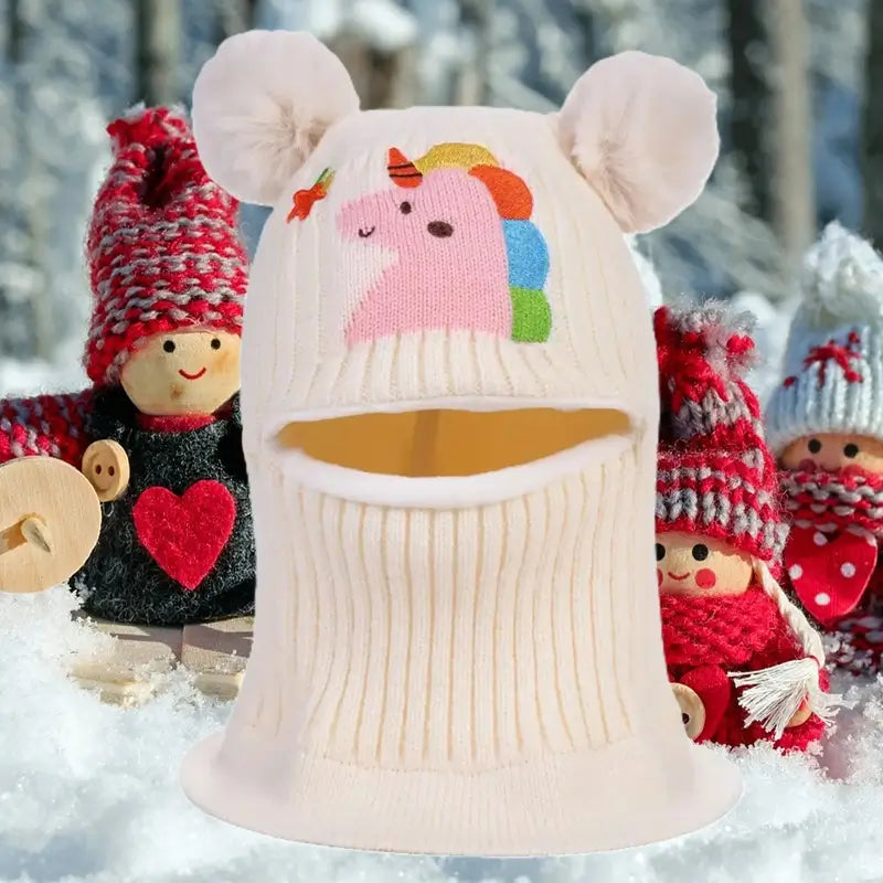 Cagoule enfant 4 ans blanche en tricot avec des oreilles pompons et motif licorne, posée dans un décor enneigé avec des poupées en arrière-plan. idéale pour l'hiver.