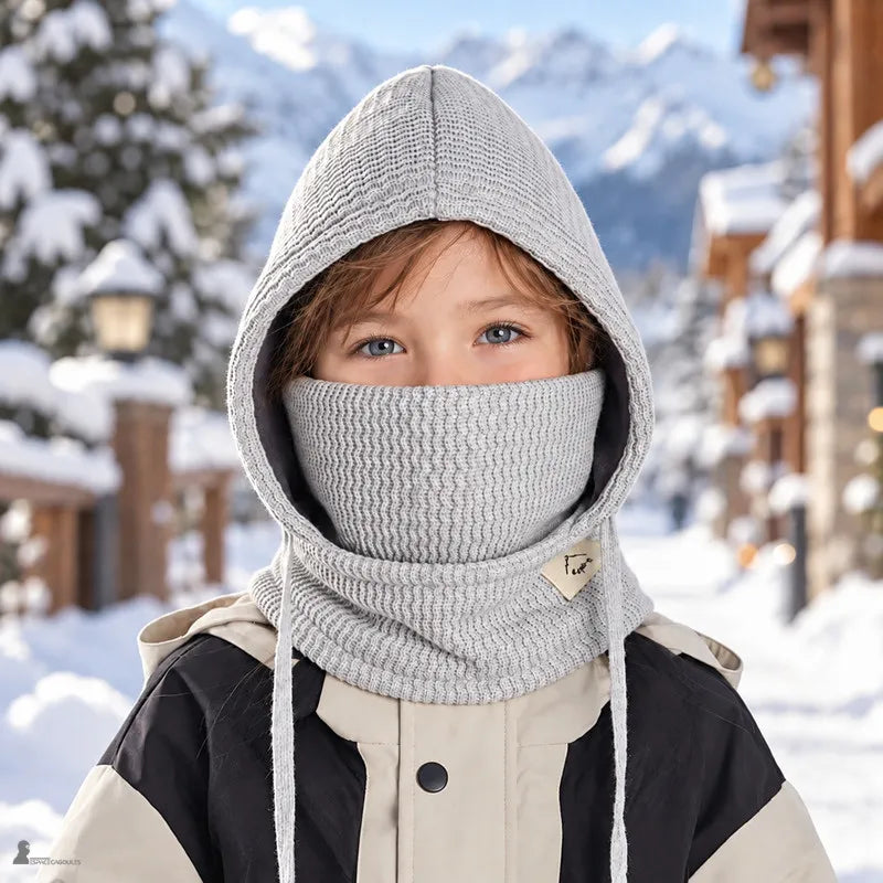 Enfant portant la cagoule hiver gris clair, capuche relevée et masque sur le nez, dans une rue de village de montagne enneigé - Espace Cagoules
