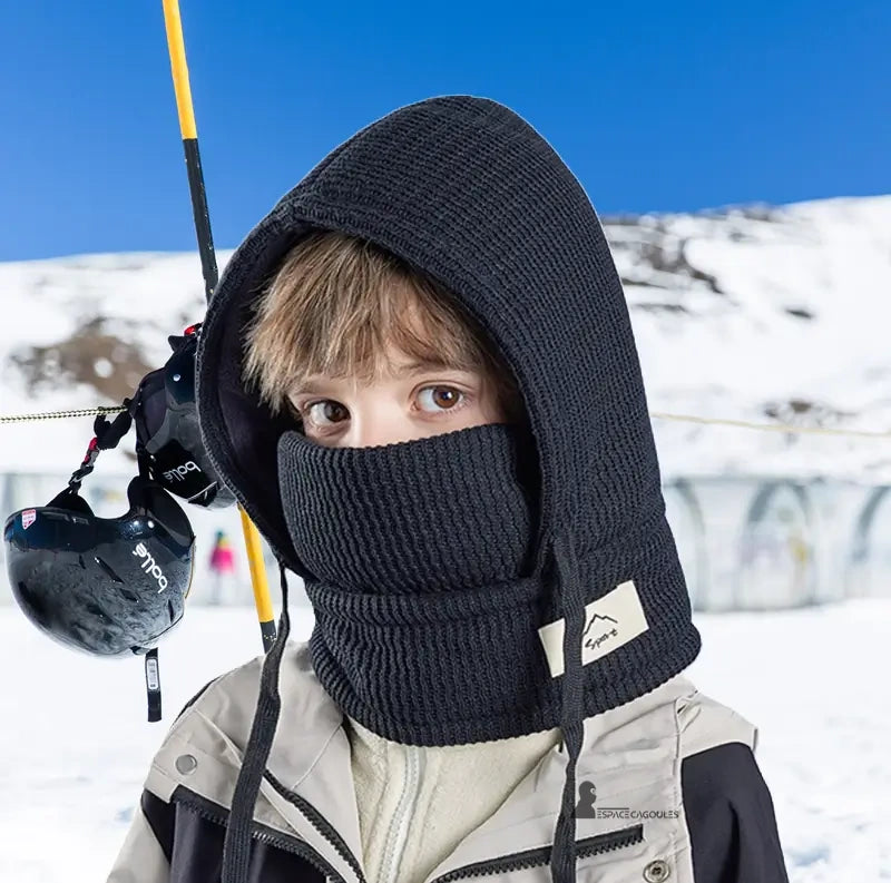 Cagoule enfant hiver avec tour de cou noire portée par un garçon en veste de ski claire dans un décor de montagne enneigée