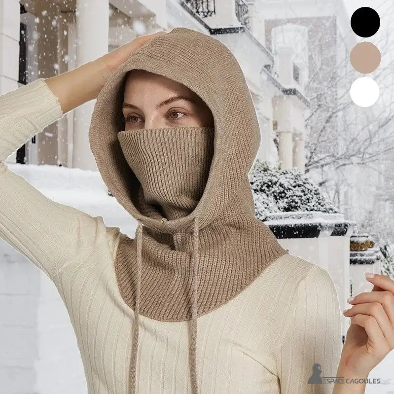 Cagoule Femme avec tour de cou intégré en maille beige, couvrant la tête et le cou, portée en extérieur sous la neige devant une maison élégante