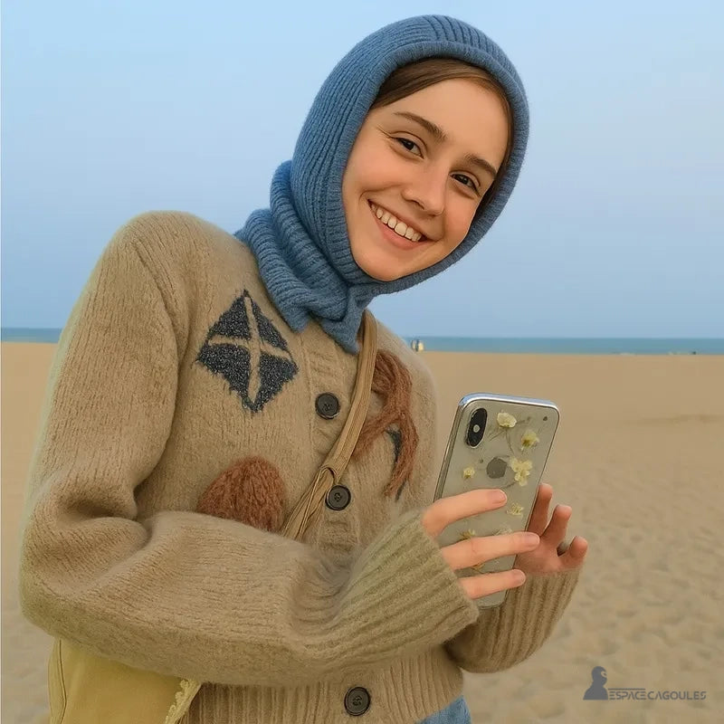 Trendige Damen-Sturmhaube, die von einer lächelnden jungen Frau am Strand getragen wird, kombiniert mit einer bestickten beigen Strickjacke, eine weiche und lässige Atmosphäre