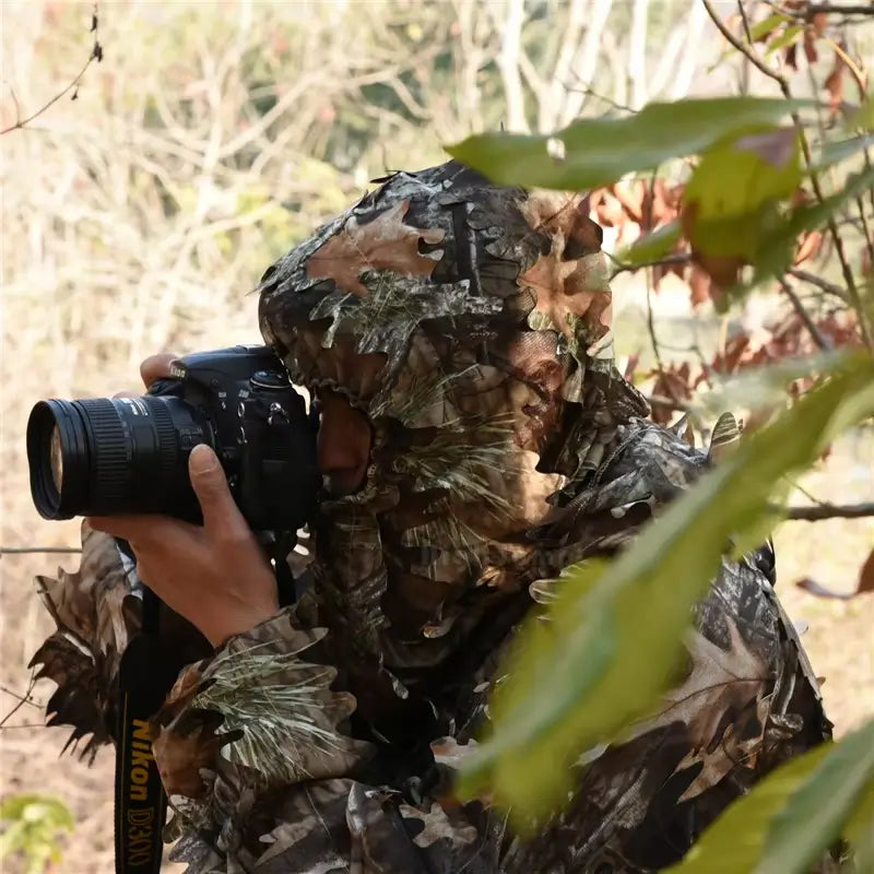 Person wearing a camouflage mesh hood in a natural environment, holding a SLR camera to photograph wildlife. The camouflage mesh balaclava offers optimal discretion for hunting or outdoor observation activities.