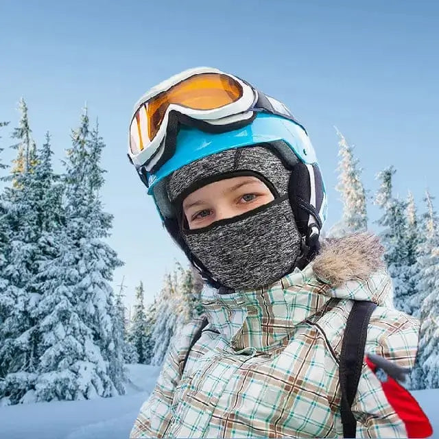 Kind, das während eines Skiausflugs eine dünne Sturmhaube unter einem grauen Kinderhelm trägt, mit schneebedeckten Bäumen und klarem Himmel im Hintergrund.