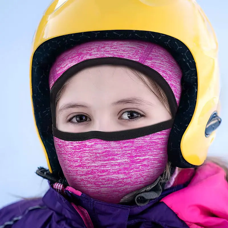 Enfant portant une cagoule fine sous casque enfant rose sous un casque jaune, prêt pour une activité extérieure hivernale.