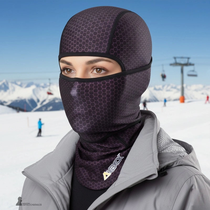 Cagoule fine sous casque en tissu technique ajusté, forme couvrante avec ouverture pour les yeux, vue portée par une femme en environnement montagne enneigé avec remontées mécaniques visibles - Espace Cagoules