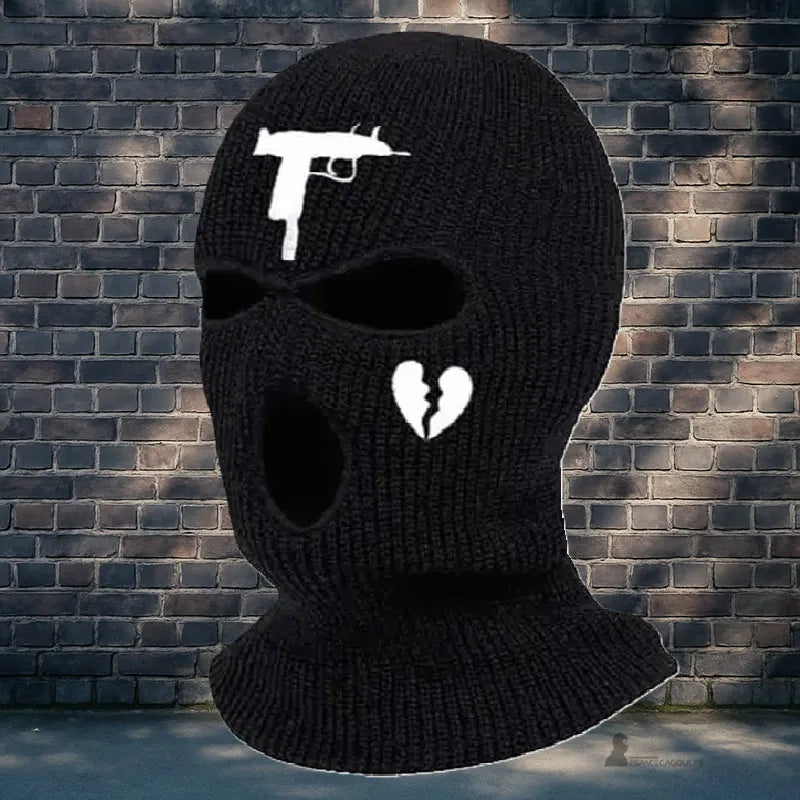 Cagoule gangster noire tricotée avec motif pistolet et cœur brisé, posée devant un mur de briques en extérieur urbain