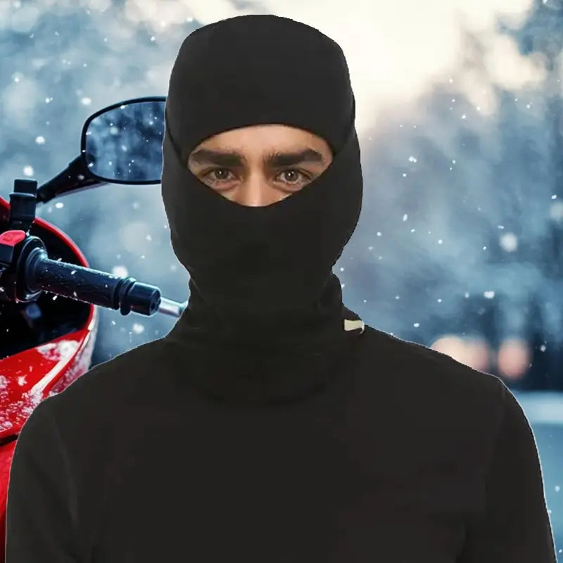 Motard portant une cagoule en laine mérinos noire, debout devant une moto dans un environnement hivernal enneigé, symbole de chaleur et de protection.