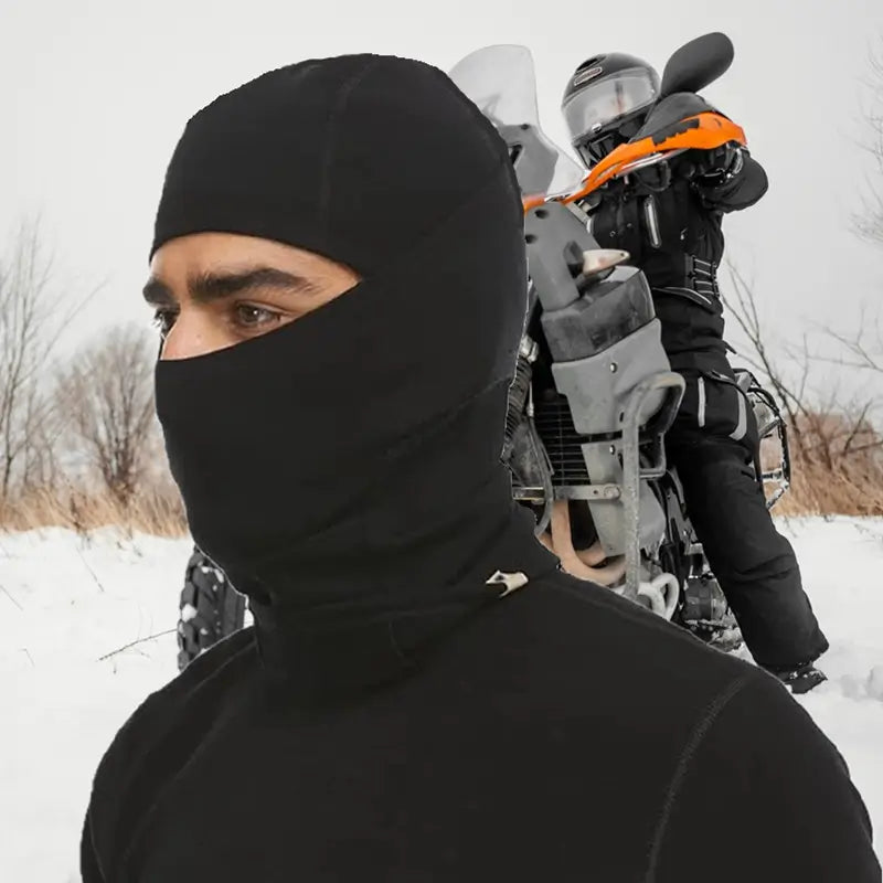 Motard portant une cagoule en laine mérinos noire, en plein air avec un équipement de ski sur l'épaule, parfait pour une protection contre le froid et le vent en hiver.