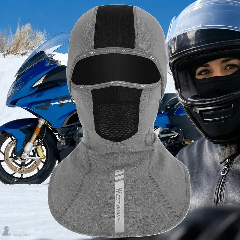 Cagoule Moto pour femme à cheveux longs en gris, conçue pour protéger du vent et du froid, portée avec un casque moto sur un arrière-plan de paysage enneigé.