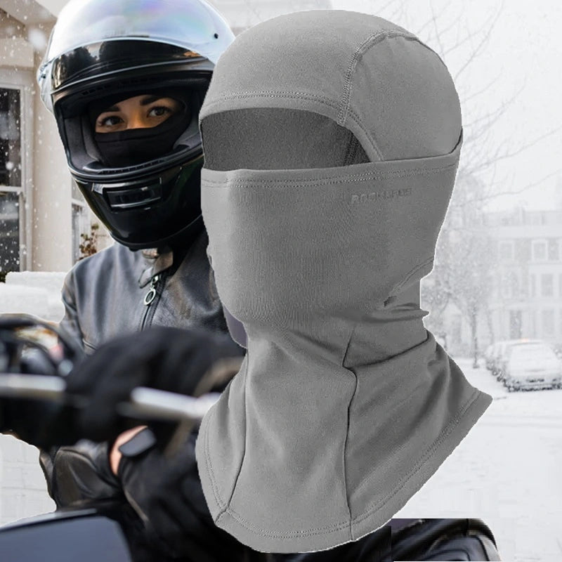 Graue Motorrad-Sturmhaube für Damen, die Gesicht und Hals vor Kälte schützt, ideal für Motorradfahrten im Winter. Im Hintergrund ein Biker, der ausgerüstet und bereit ist, im Schnee zu fahren.