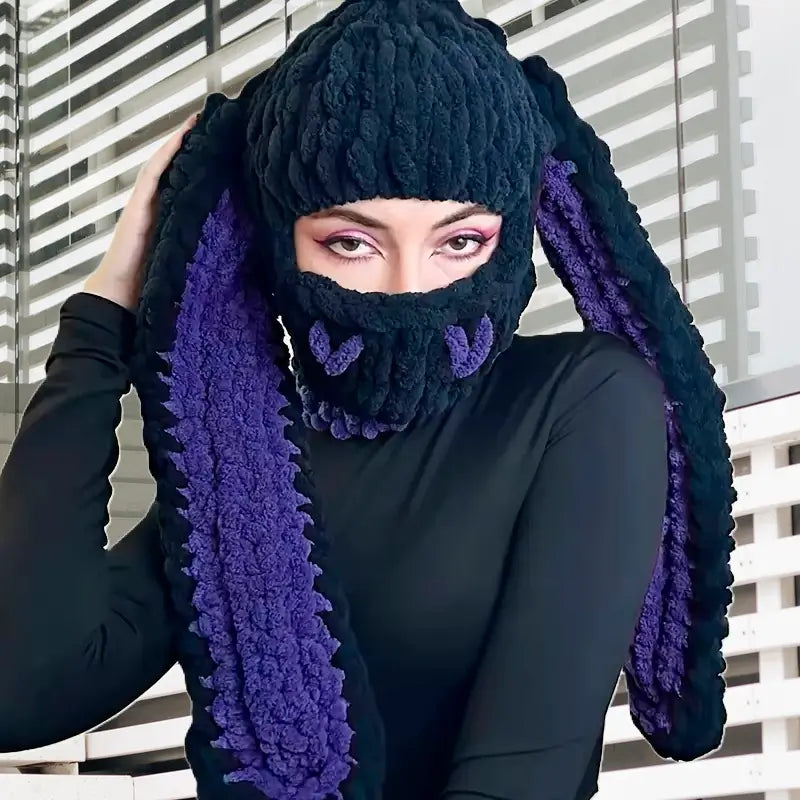 Balaclava Black and purple rabbit ears â A woman in black outfit wears a black knitted balaclava with purple drooping rabbit ears. It is indoors with a modern background.