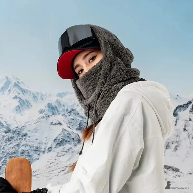 Cagoule polaire Femme grise portée par une femme avec casquette rouge et lunettes de ski, arrière-plan de montagnes enneigées