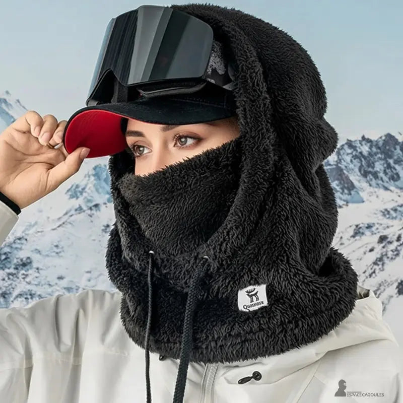 Cagoule polaire Femme noire épaisse portée avec casquette rouge et masque de ski, photographie en montagne enneigée