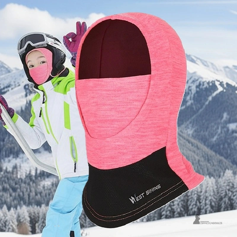 Rosa skidbalaklava för tjejer som bärs i backen av en ung skidåkare i gröna och blå kläder, som kombinerar värme, komfort och skydd under hjälmen – Espace Cagoules