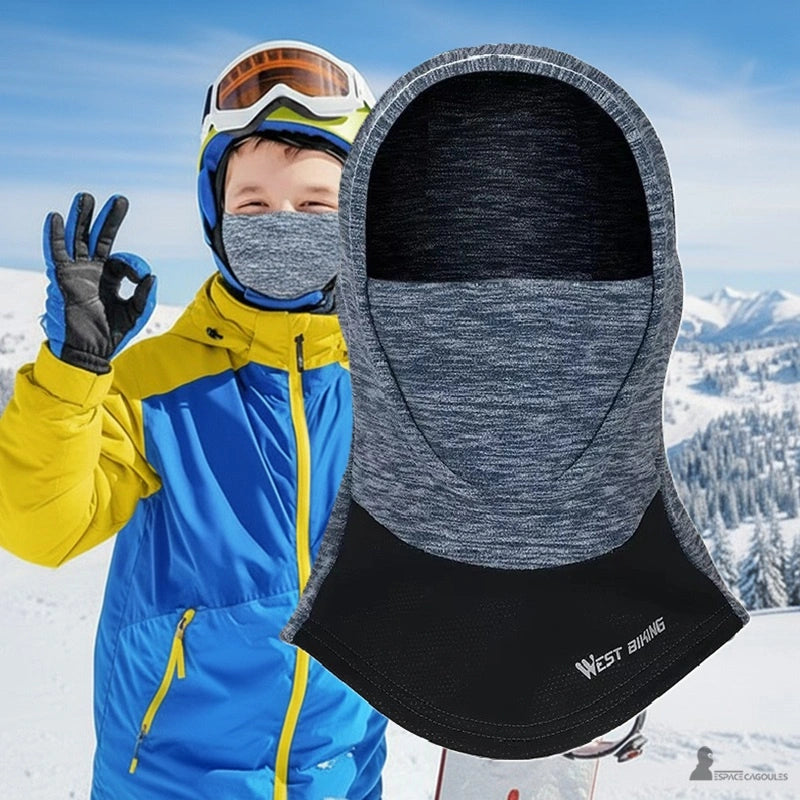 Grå skidbalaklava för pojkar som bärs i de snöiga backarna av ett barn i blå och gula skidkläder, som erbjuder värme och skydd under hjälmen – Espace Cagoules