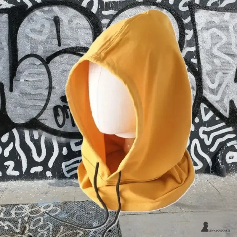 Cagoule streetwear jaune moutarde en coton, posée sur un mannequin blanc devant un mur de graffitis urbains en noir et blanc, accessoire mode tendance pour look décontracté