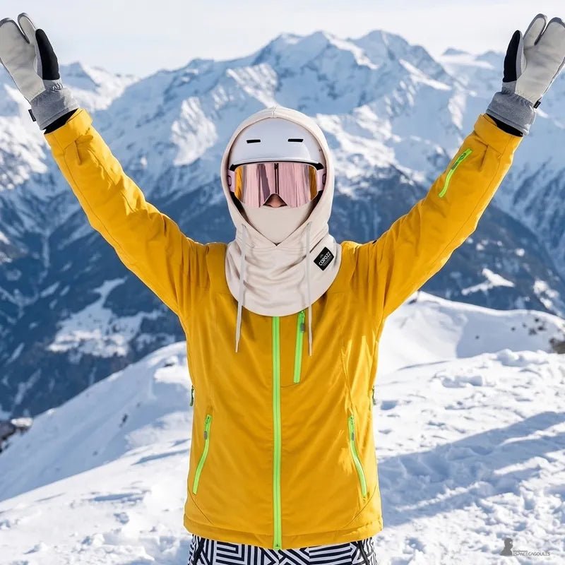 Cagoule sur casque de ski crème portée avec casque et masque par une skieuse bras levés devant un panorama de montagnes enneigées - Espace Cagoules