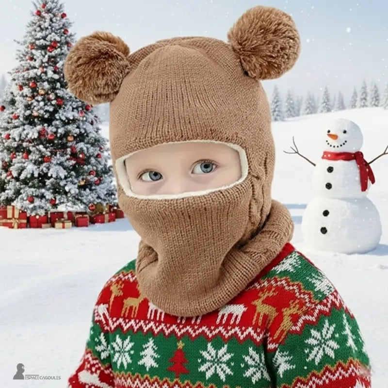 Cagoule tricot enfant marron remontée couvrant le visage, petit garçon en extérieur dans la neige devant un sapin et un bonhomme de neige