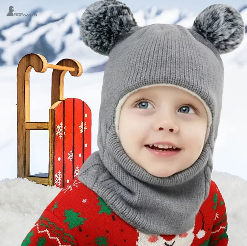 Cagoule tricot enfant grise avec pompons, petit garçon souriant en extĂ©rieur dans la neige Ă cĂŽtĂ© dâune luge rouge