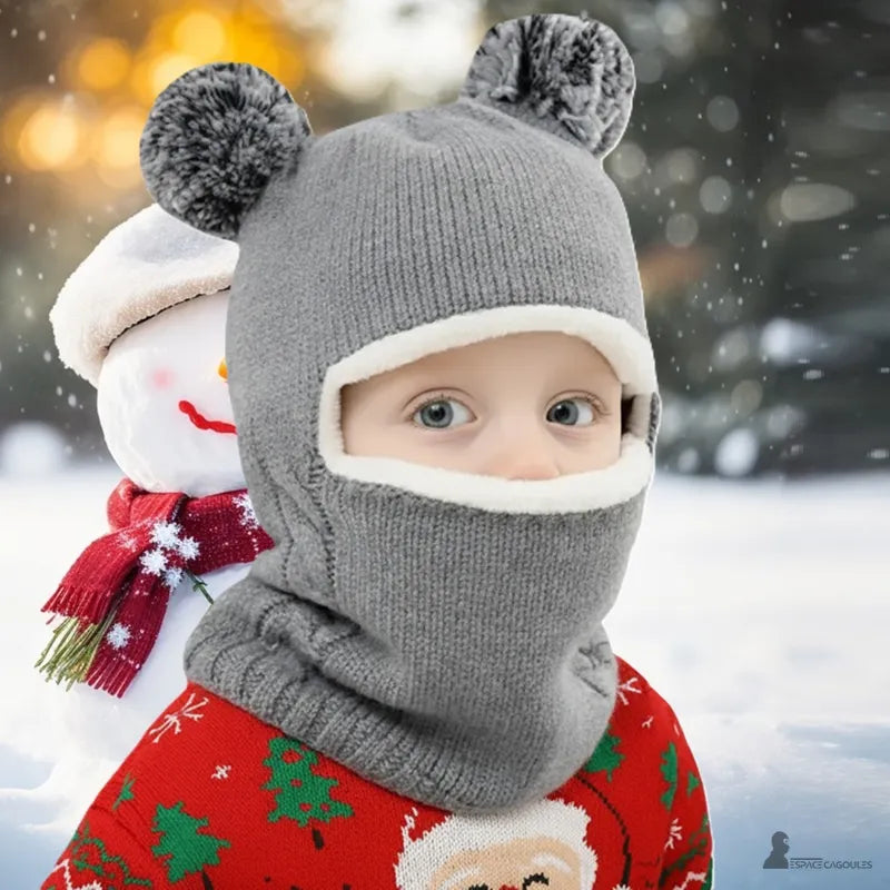 Cagoule tricot enfant grise remontée sur le visage avec pompons, petit garçon en extérieur devant un bonhomme de neige